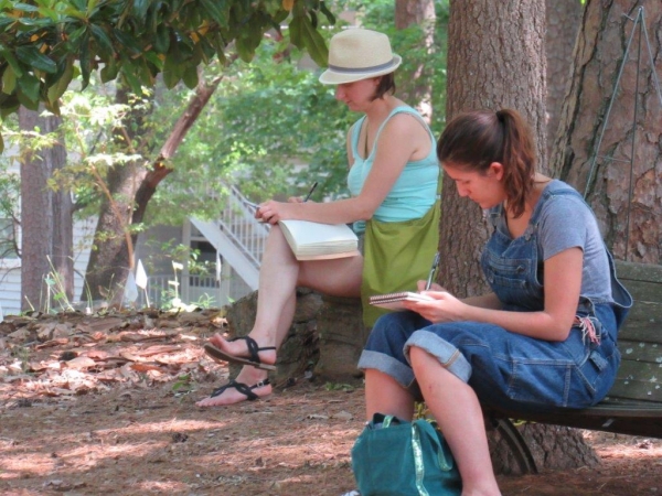 Two visitors sitting under trees and writing in notebooks at the Celebrate The "O" BBQ event.